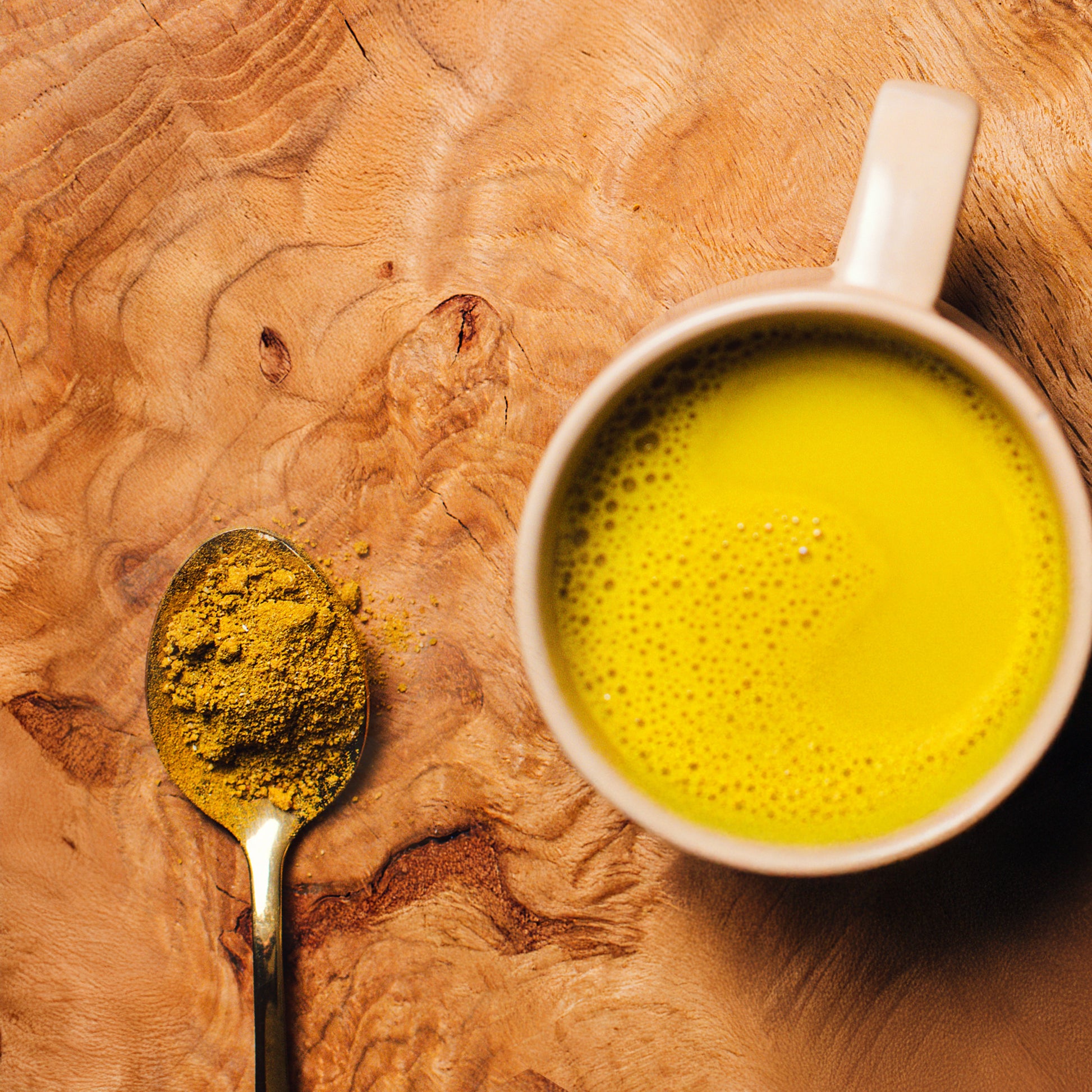 Mug of Turmeric Milk mix on a wooden surface with a spoonful of drink mix powder on wood surface.